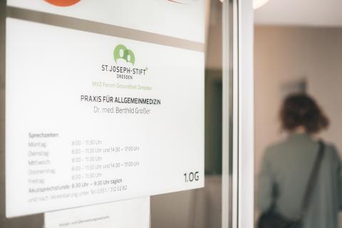 Praxis für Allgemeinmedizin, Eingangsbereich der Praxis mit Türschild und Sprechstundenzeiten im Vordergrund.